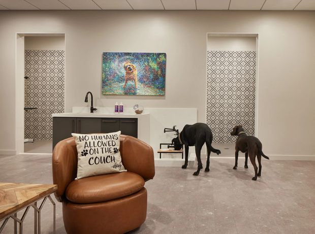 A warm, contemporary open-plan interior featuring a compact kitchenette or bar with dark cabinetry and a pale benchtop, flanked by two recessed decorative screens with geometric patterned panels. A leather lounge chair and coffee table occupy the foreground, while sculptural animal figures and colourful artwork add visual interest. The room has a smooth pale concrete-like floor, neutral walls, and a suspended grid ceiling.