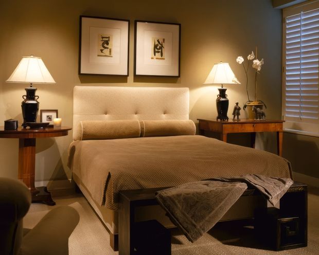 A warmly lit, refined bedroom centered on an upholstered king-size bed with a tall tufted cream headboard, cylindrical bolster cushions, and textured taupe bedding. Matching dark wood bedside tables hold traditional lamps and decorative objects, while framed artwork, an orchid arrangement, and white plantation shutters complete the elegant interior.