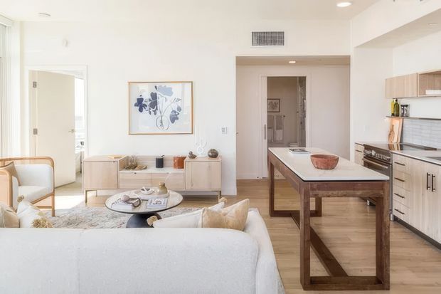 Bright open-plan living and kitchen interior with a white upholstered sofa in the foreground, a round coffee table and low timber media console, and a timber kitchen island with a pale benchtop. Light timber flooring, white walls, pale cabinetry, recessed lighting, and framed botanical artwork create a clean, warm, contemporary feel.