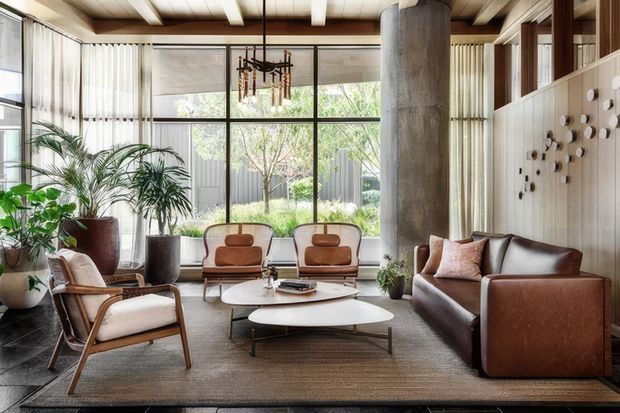 A warm, mid-century-inspired living room with expansive floor-to-ceiling glazing, sheer curtains, timber-lined ceiling, and a prominent cylindrical concrete column. The room features a brown leather sofa, sculptural upholstered lounge chairs, layered oval coffee tables, indoor palms, a textured rug, and decorative wall art. The setting is bright and connected to a landscaped courtyard or garden outside.