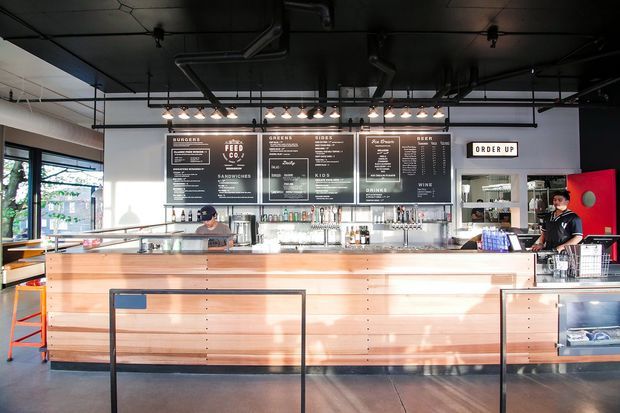 Bright contemporary commercial food-service interior showing a long timber-fronted service counter and bar, illuminated menu boards, beverage taps, display shelving, and staff behind the counter. The space has an exposed dark ceiling with visible ductwork and track-style pendant lighting, polished concrete flooring, and large glazed openings on the left.