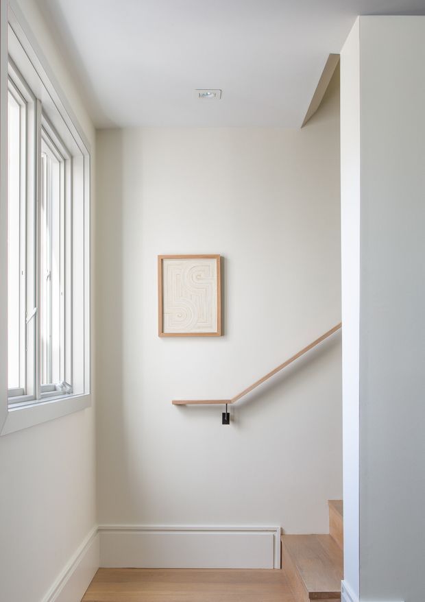 Bright, minimalist stairwell with a large white-framed window, pale painted walls, light timber flooring and a simple timber handrail with black metal supports. A small framed abstract artwork provides a subtle decorative feature, while a recessed ceiling light and clean trim reinforce the understated design.