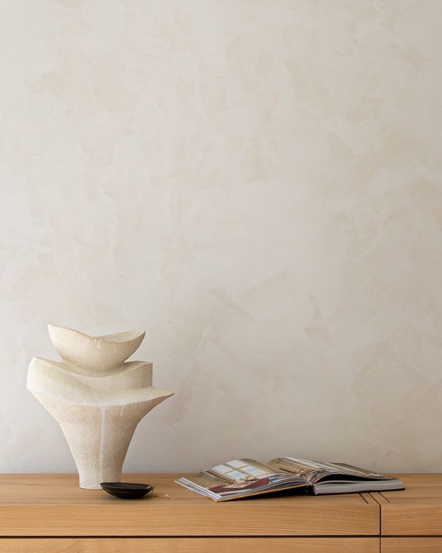 A softly lit interior vignette featuring a warm light-wood sideboard with clean horizontal drawer fronts, an open design magazine, and sculptural cream ceramic vessels. The background wall has a subtle cloudy, hand-trowelled plaster texture, creating a calm, minimalist composition.