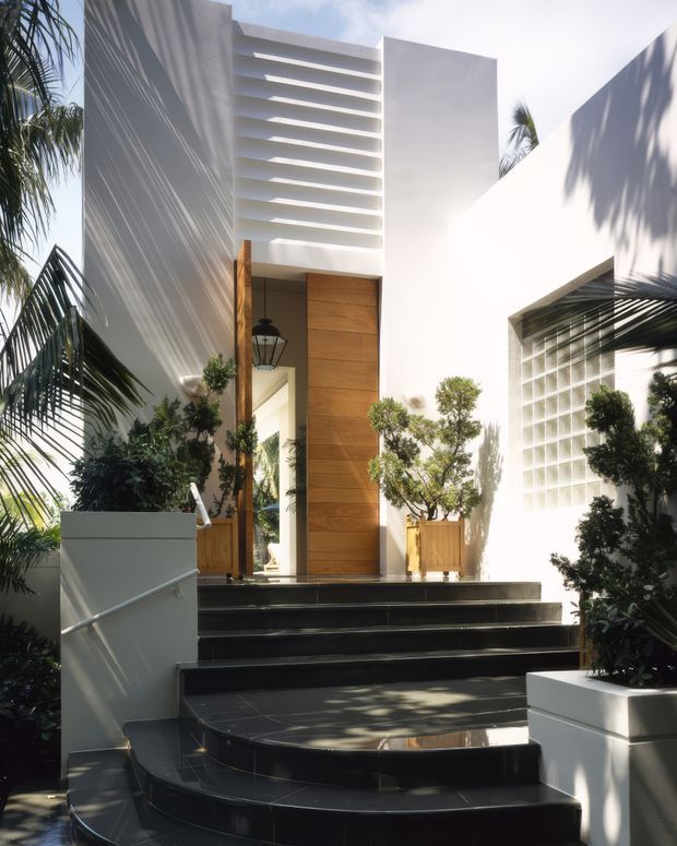 A bright contemporary residential entrance with broad black tiled steps leading to a tall timber pivot door set within a white rendered facade. Horizontal louvres above the entry and a glass-block feature wall add texture, while tropical planting and strong palm shadows frame the approach.