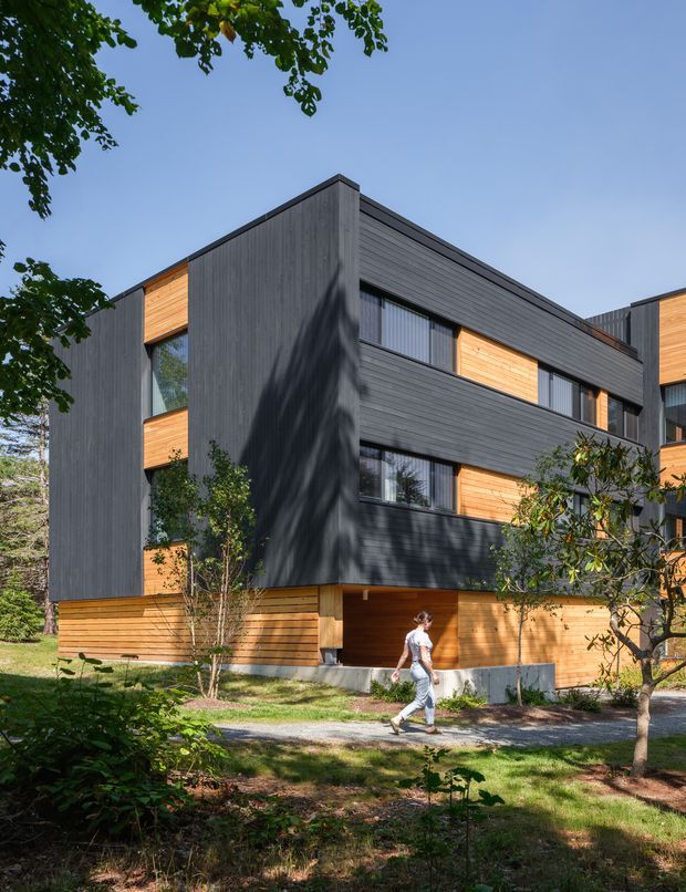Exterior architectural photograph of a contemporary multi-storey building set among mature trees and landscaped grounds. The facade combines dark vertically textured cladding with warm horizontal timber cladding, large rectangular windows, projecting box-like volumes, and clean flat rooflines. A pedestrian is visible in the foreground.