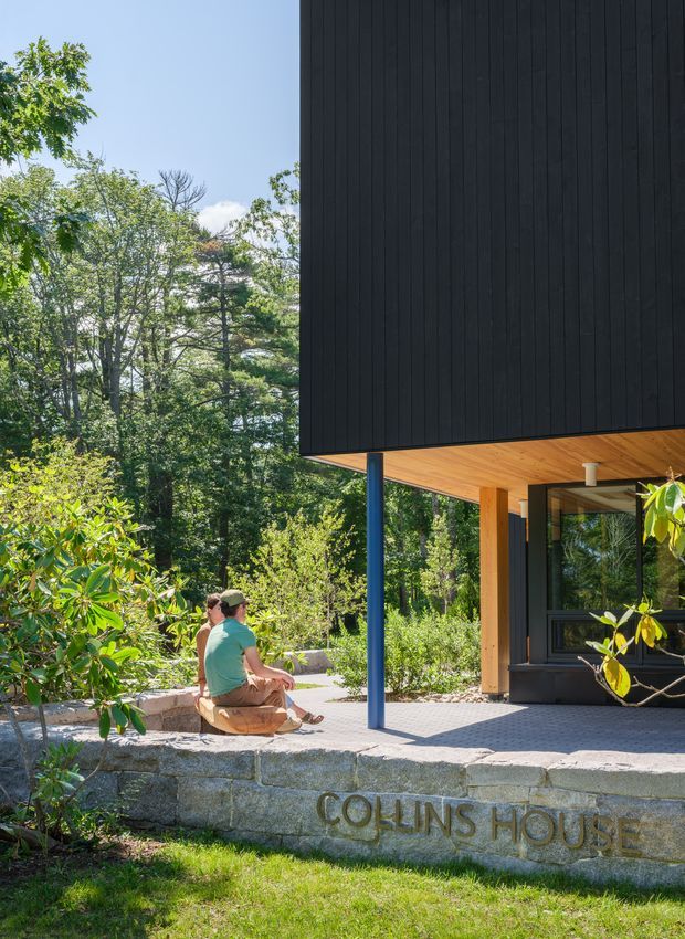 Exterior view of a contemporary house set within a leafy, wooded landscape. The dark vertical-clad upper volume projects over a covered entry terrace, supported by a slender blue post, with warm timber soffit lining and timber-framed glazing below. A paved terrace, low stone retaining wall engraved “COLLINS HOUSE,” native planting and seated people establish the residential entrance setting.