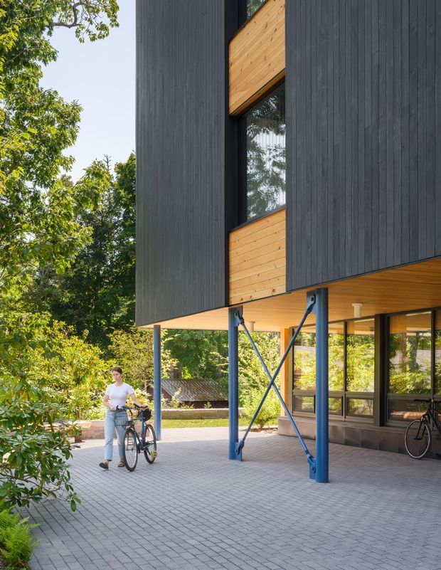 Exterior architectural photograph of a contemporary elevated building in a leafy setting. The structure features dark vertical timber cladding, warm timber soffits and inset panels, extensive glazing with dark frames, and exposed blue-painted steel support columns with diagonal bracing. A paved covered forecourt sits beneath the cantilevered upper volume, with bicycles and a person visible.