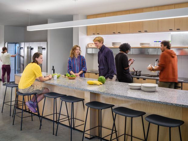 A bright, spacious communal kitchen with a long central island, integrated timber cabinetry, open shelving, stools, and several people preparing food. The room has a clean contemporary fit-out with a grey stone-like benchtop, pale timber joinery, white walls, and a polished concrete floor.