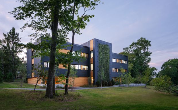 Exterior view of a contemporary multi-storey building set within a mature landscaped site. The building has dark grey upper cladding, warm timber-clad lower walls, extensive rectangular glazing, projecting box-like volumes, and a tall central facade element partly covered with climbing greenery. Large established trees and open lawn surround the structure.