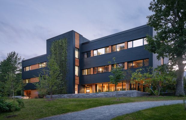 A contemporary multi-storey building viewed from landscaped grounds at dusk. The dark horizontal cladding, crisp rectangular massing, extensive window bands, and warm timber-toned recessed sections create a modern institutional or commercial facade. Mature trees, lawn, stone retaining elements, and a curved gravel or paved approach are visible in the foreground.
