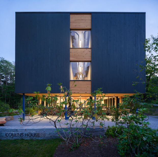 Front exterior view of a contemporary multi-storey house with a tall, simple rectangular form. The upper levels are clad in dark blue-black vertical boards, with a central vertical strip of warm timber cladding and recessed windows. The ground floor is largely glazed and set beneath the upper volume, creating a sheltered, open entry or living zone. Landscaping, bicycles, paving, and trees surround the building.