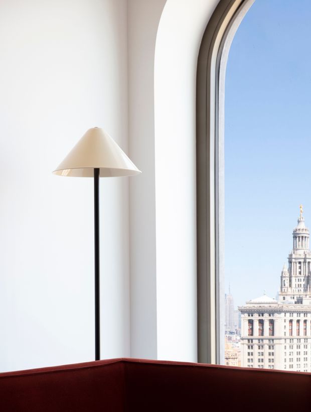 Minimal, bright interior corner with a tall black floor lamp and cream conical shade beside a large arched window. A deep burgundy upholstered sofa or chair occupies the foreground, while an ornate urban tower is visible through the window.