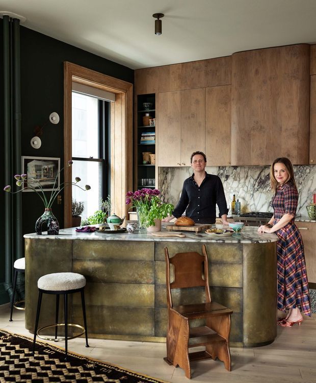 A richly styled kitchen centered on a large island with a veined stone benchtop and aged brass or bronze-toned panelled cabinetry. Warm timber wall cabinetry, a dramatic dark green wall, timber-framed window, marble-look splashback, open shelving, stools, and a distinctive wooden chair create an eclectic, vintage-influenced interior. Two people stand behind the island, with plants, flowers, cookware, and food adding a lived-in atmosphere.