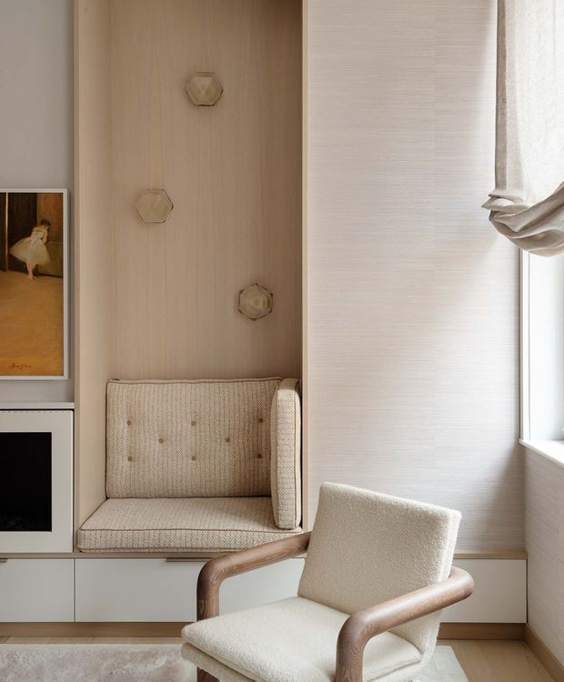 A warm, refined sitting nook with a built-in upholstered bench set into a light timber wall, paired with a cream boucle armchair with curved timber arms. A small fireplace or dark display recess and framed artwork sit to the left, while a softly draped window and pale textured wall covering brighten the space. The palette is predominantly cream, beige, and light wood with subtle sculptural wall lighting.
