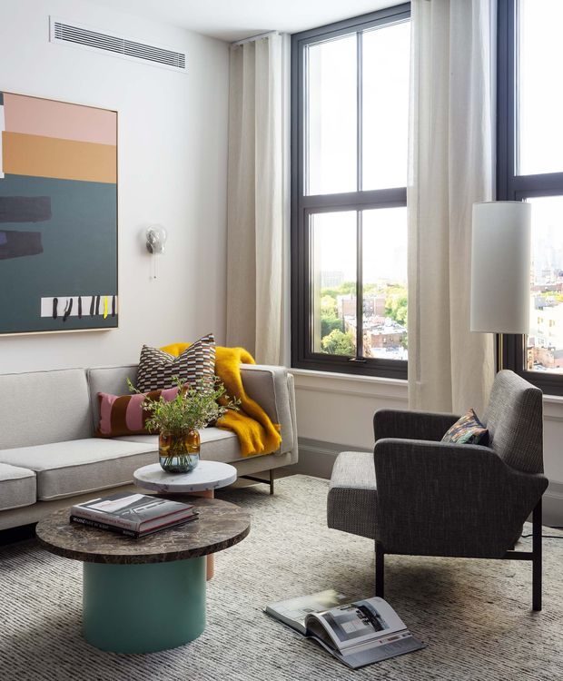Bright, refined living room with floor-to-ceiling black-framed windows overlooking an urban setting. The room features a light grey sofa, dark upholstered armchair, layered round coffee tables, a sculptural wall sconce, geometric abstract artwork, patterned cushions, and a mustard throw. Neutral curtains and a textured pale rug soften the contemporary interior.