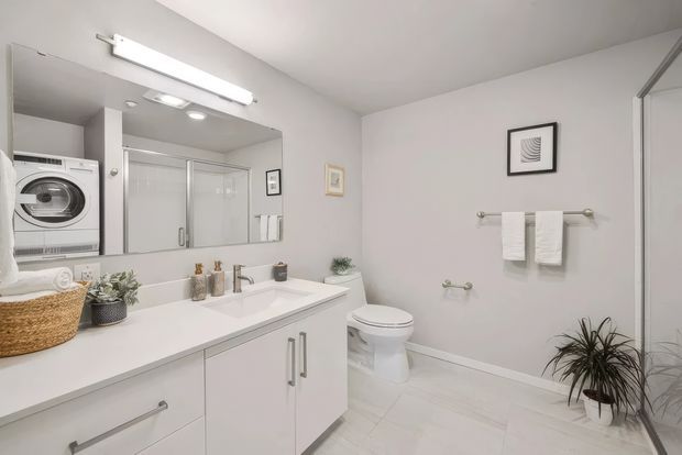 Bright, clean contemporary bathroom with a large mirrored vanity, white cabinetry and benchtop, integrated double basin, toilet, glass-enclosed shower, wall-mounted towel rails, and pale tiled flooring. A stacked washer and dryer are visible in a recessed area at left, suggesting a combined bathroom-laundry space. The room is styled in a minimal neutral palette with a few plants and framed artwork.