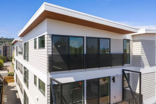 Exterior architectural view of a contemporary multi-storey residential building with white horizontal cladding, broad flat roof overhangs, warm timber soffits, dark-framed glazing, and black horizontal balcony privacy screens and balustrades. The image focuses on the upper-level facade and balconies in bright daylight.