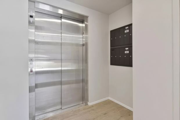 A compact apartment building lift lobby with stainless-steel elevator doors and control panel, dark wall-mounted mailboxes, white painted walls, and light timber-look flooring. The space is clean, functional, and minimally furnished.