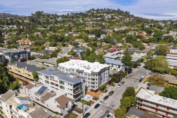 Aerial view of a dense suburban urban neighbourhood with a prominent multi-storey white apartment or mixed-use building in the foreground, surrounded by residential homes, streets, mature trees, and a hilly built-up landscape. The scene suggests a developed suburban setting with low- and mid-rise buildings and distant water or coastline visible at the horizon.