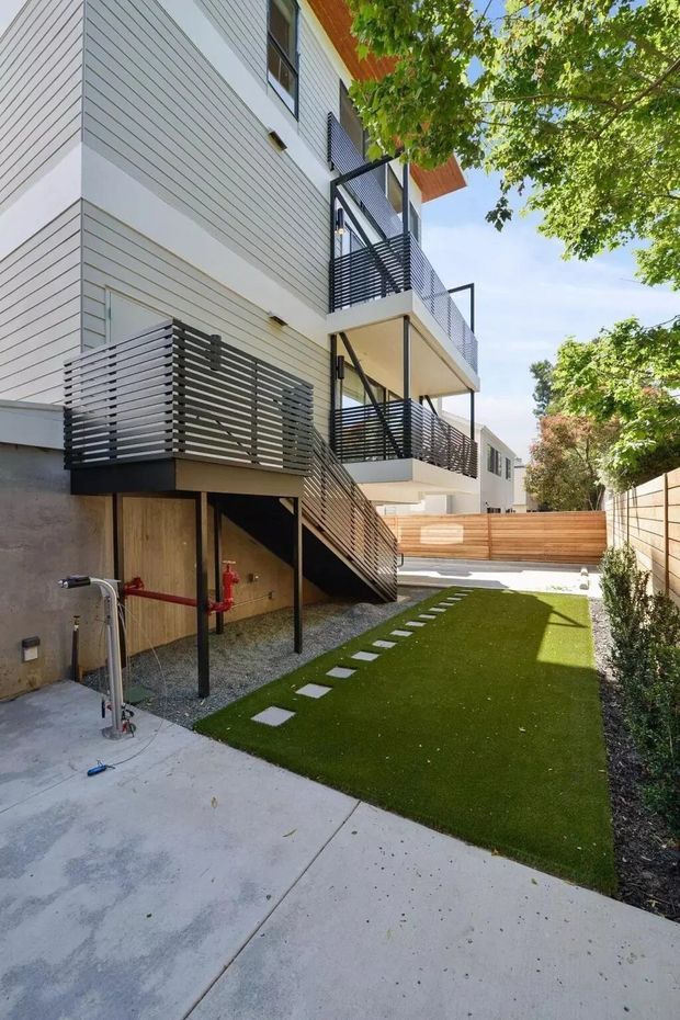 Exterior view of a contemporary multi-storey residential building with pale horizontal cladding, projecting balconies, dark horizontal metal privacy screens and railings, and an external stair. The building sits beside a narrow landscaped courtyard with artificial turf, stepping-stone pavers, concrete paving, timber fencing, and mature trees.