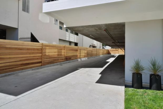 A contemporary multi-unit residential exterior showing a paved driveway or access lane beneath a large concrete overhang. The scene includes smooth white rendered walls, a horizontal timber privacy fence, dark asphalt paving, concrete surfaces, and tall black planters with ornamental grasses. The image is bright and clean, with strong linear geometry and covered vehicle-access space.