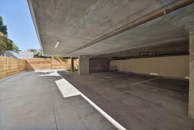 A covered open-air concrete parking area beneath a building, with multiple marked parking bays, exposed concrete slab ceilings, structural columns, overhead utility piping, and a timber privacy fence along the paved driveway or access area. Bright daylight enters from the open side.