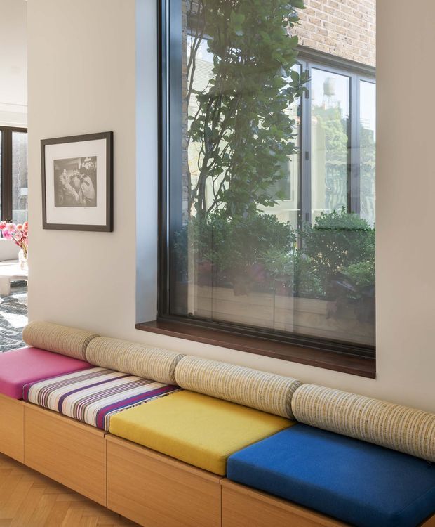 A bright contemporary interior corner featuring a long built-in window seat beneath a large dark-framed window. The seat has integrated warm timber cabinetry and vividly coloured upholstered cushions in yellow, blue, pink, and striped fabric, with a cylindrical textured bolster along the back. A framed monochrome artwork hangs on the pale wall, while greenery and a brick exterior courtyard are visible through the glazing.