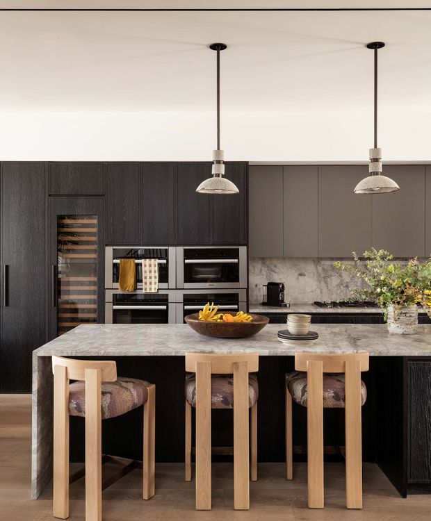 A refined contemporary kitchen centred on a large marble island with integrated seating for four. The kitchen features dark vertically grained cabinetry, built-in black appliances, a grey stone backsplash and benchtop, warm timber stools with patterned upholstered seats, and two pendant lights. The palette is sophisticated and warm, combining charcoal cabinetry, pale stone and natural wood.