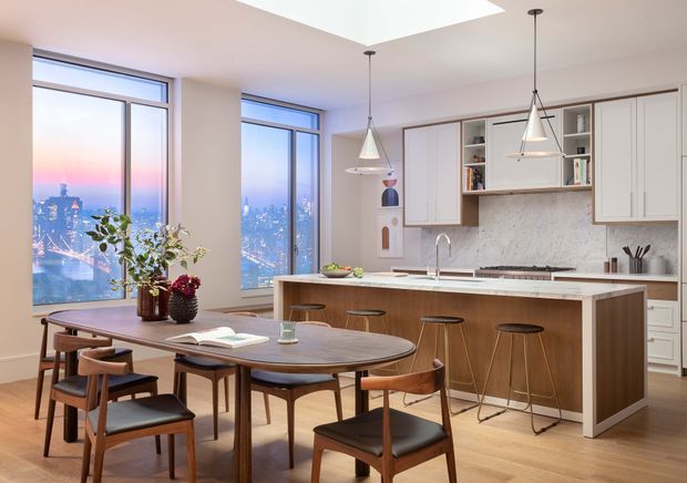 Bright contemporary open-plan kitchen and dining area in a high-rise setting, with large floor-to-ceiling windows overlooking a city skyline. The room features a long timber island with a pale stone benchtop, integrated cabinetry, pendant lights, a marble-look splashback, and a rounded timber dining table with mid-century-inspired chairs.