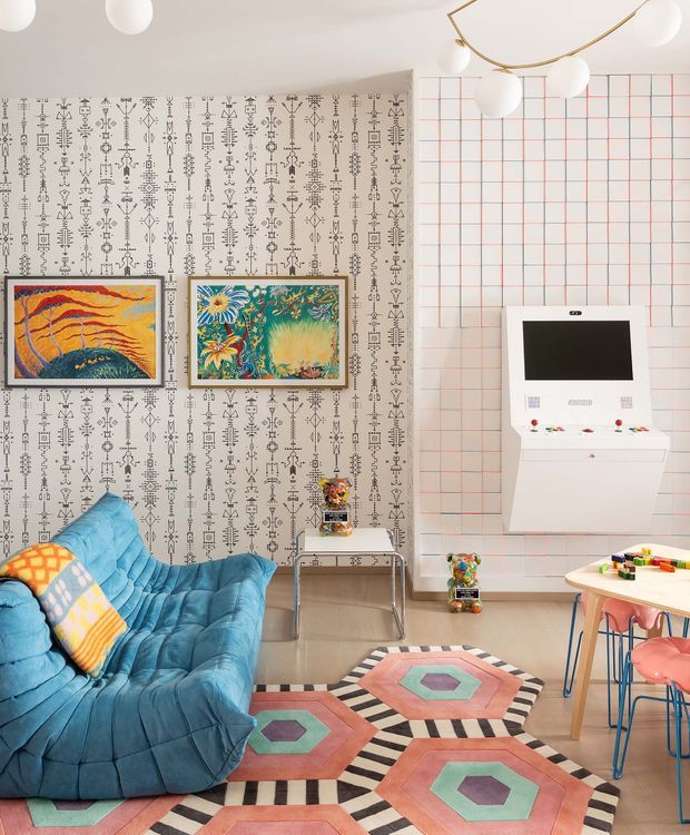 A bright, eclectic play and living space with a deep blue tufted sectional sofa, colourful geometric rug, patterned wallpaper, framed artwork, and a wall-mounted white entertainment or storage unit. The right side includes a small children's table with colourful chairs and toys. The ceiling has sculptural white globe pendant lights, while the walls combine a black-and-white graphic pattern with a red grid motif.