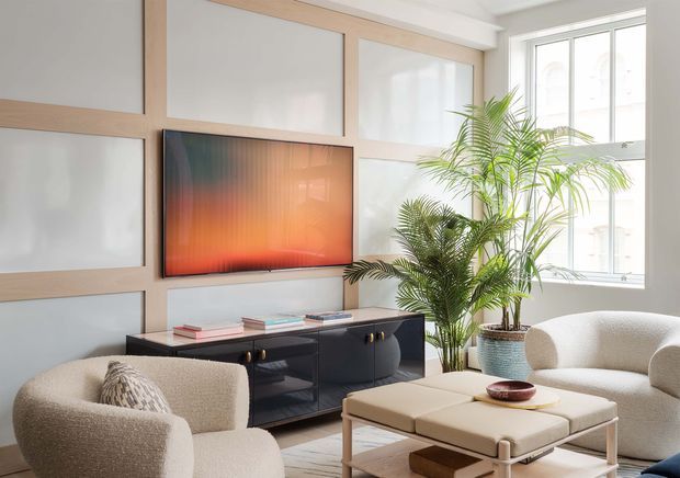 Bright contemporary living room with a cream boucle lounge chair and sofa, a pale upholstered ottoman, and a dark low media console. The room features a large abstract wall-mounted television or artwork, light timber-framed frosted wall panels, tall windows, and indoor palm plants. The palette is soft and neutral with warm timber accents and a vivid orange-red screen accent.