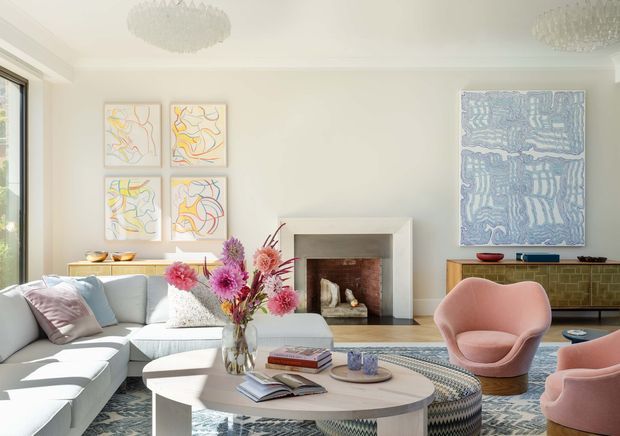 Bright, refined living room with a large pale sectional sofa, sculptural blush-pink lounge chairs, a rounded coffee table, patterned rug, colourful floral arrangement, contemporary artwork, and a central white fireplace with a timber-toned interior. Large windows bring in abundant natural light, while crystal-style ceiling fixtures add a decorative accent.
