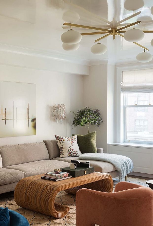 A refined, light-filled living room with a curved beige sectional sofa, sculptural ribbed wood coffee table, rust-colored lounge chair, patterned cushions, and a soft blue throw. The room features white walls and ceiling mouldings, a large window with a Roman shade, abstract artwork, greenery, and a distinctive brass multi-arm pendant light with opal globes. The overall look is warm, elegant, and contemporary with organic curved forms.