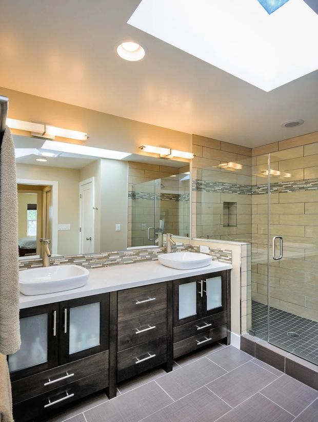 Bright contemporary bathroom with a large double vanity, two vessel sinks, dark wood cabinetry, a tiled backsplash, and a spacious glass-enclosed walk-in shower. The room features beige wall tiles with a horizontal mosaic accent band, grey floor tiles, mirrored cabinetry, and recessed and wall-mounted lighting.
