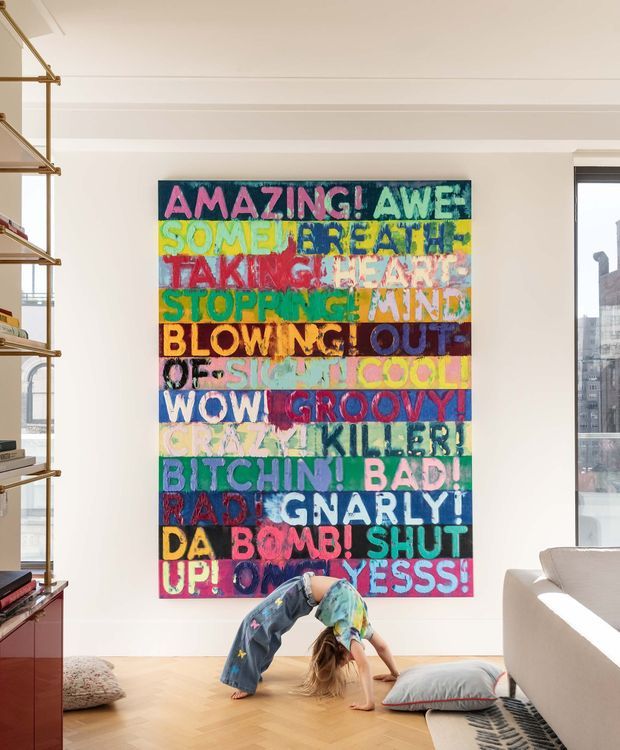 Bright contemporary living room with white walls, decorative ceiling moulding, pale timber flooring, a large colourful typographic artwork, open shelving with brass-toned supports, and a light upholstered sofa. A child is playfully posing on the floor, with cushions and a partially visible rug adding casual warmth. Tall glazing at the right brings in daylight and shows an urban exterior.