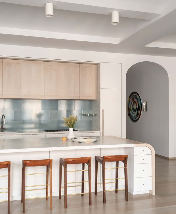 A bright, refined contemporary kitchen featuring a large white island with rounded waterfall-style end, integrated white cabinetry, pale timber overhead cupboards, and a glossy blue-grey tiled splashback. Four warm timber and leather bar stools with brass foot rails line the island. A sculptural arched wall opening leads to a hallway, with recessed tray ceiling detailing and simple cylindrical pendant fixtures overhead.