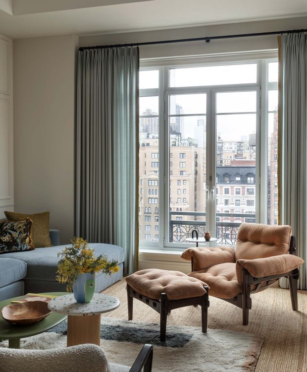 A refined living room in a high-rise apartment, centered on a large multi-pane window with city views and dark full-length curtains. The room features a pale blue sectional sofa, a tan upholstered lounge chair with matching ottoman, layered area rugs, sculptural nesting coffee tables, and a small floral arrangement. Light walls, generous glazing, and softly textured furnishings create a bright yet warm, contemporary setting.
