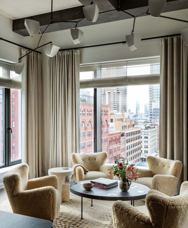 A refined high-rise living room with expansive corner windows overlooking a dense city skyline. The room features cream floor-to-ceiling curtains, sculptural pendant lights suspended from dark exposed ceiling beams, several plush boucle lounge chairs, a round black coffee table, a geometric area rug, and a small side table. The palette is warm neutral with a sophisticated contemporary character.