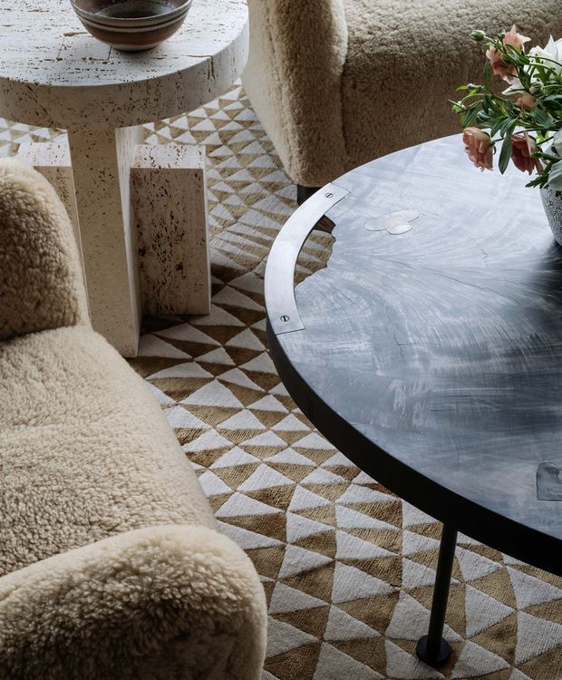 Close-up interior scene featuring a round dark stone coffee table with a curved metal inlay, sculptural light stone side table, and plush cream shearling-style seating. A geometric patterned rug in cream, taupe, and brown grounds the composition, with a floral arrangement adding a soft natural accent. The room has a tactile, luxurious, softly layered contemporary aesthetic.