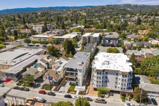 Aerial view of a suburban urban neighbourhood with several low- and mid-rise apartment and commercial buildings, streets, mature trees, and rolling hills in the background. The scene shows contemporary flat-roofed development integrated among established residential properties.
