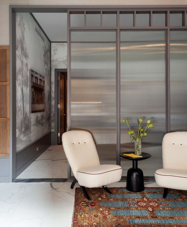 A refined interior sitting area viewed from a hallway or entry zone. Two cream upholstered lounge chairs sit on a richly patterned Persian-style rug, paired with a small black sculptural side table, vase, and yellow flowers. Behind them is a dark-framed partition with ribbed translucent glass panels and upper clear panes. The adjacent hall features pale marble-look flooring, dark wall panelling, a large monochrome mural or textured wall finish, and timber detailing. The overall composition is sophisticated, warm, and contemporary with strong contrast between cream upholstery, dark framing, and colourful rug.