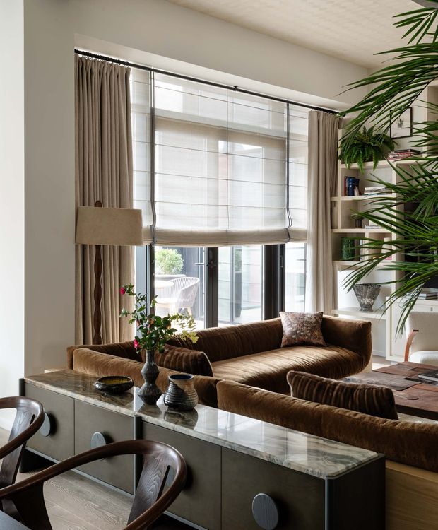 A refined living room with a warm brown velvet sectional sofa, large glazed doors and windows dressed with sheer Roman blinds and full-height taupe curtains, and a marble-topped console dividing the seating area from a dining space. Built-in open shelving, indoor palms, sculptural vases, and dark timber dining chairs add layered contemporary character. Natural daylight creates a warm, elegant atmosphere.