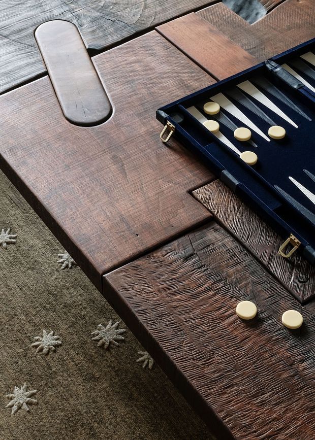 Close-up still life of a dark, richly textured timber coffee table with varied plank surfaces and a carved elongated inset detail. An open backgammon set with cream playing pieces rests on the table, which sits over a muted patterned rug. The image emphasises tactile wood grain, craftsmanship and warm, moody styling.