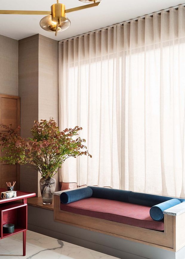 A refined living room with a built-in window seat beneath full-height sheer curtains. The space features warm taupe wall panelling, dark timber joinery, a burgundy upholstered bench cushion, cylindrical navy bolsters, a sculptural brass ceiling fan-light, and a marble-look floor. A large indoor plant and red open shelving add colour and character.