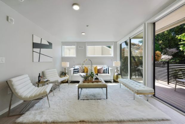 Bright contemporary living room with white upholstered lounge chairs, a sofa, glass coffee table, and a large pale rug. Full-height sliding glass doors open onto a timber deck with horizontal black screening, while large windows, recessed ceiling lights, and light grey walls create an airy indoor-outdoor connection.