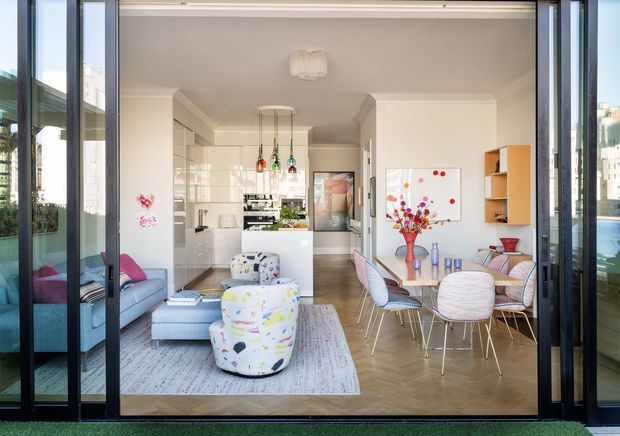 Bright open-plan apartment interior viewed through fully glazed dark-framed sliding doors, combining a compact living area, dining area, and white kitchen. The room features light timber flooring, white cabinetry, colourful pendant lights, a patterned rug, upholstered lounge seating, and a round dining table with modern chairs. Outdoor greenery and surrounding buildings are visible beyond the glazing.
