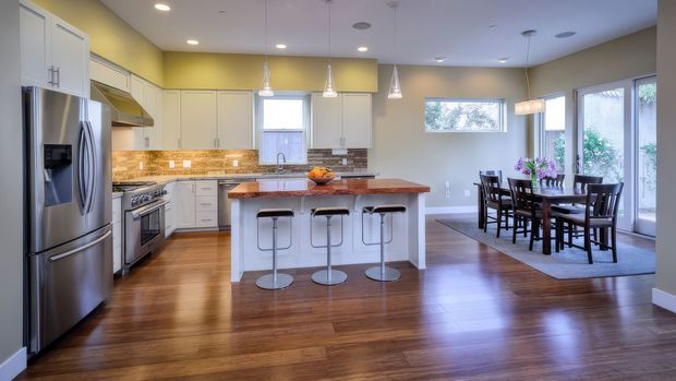Bright open-plan kitchen and dining area with white shaker-style cabinetry, stainless-steel appliances, warm wood flooring, a central island with a timber benchtop and seating for three, and a separate dark wood dining table near large glazed doors. The space has a warm yellow-green accent wall, pendant lighting, and abundant natural light.