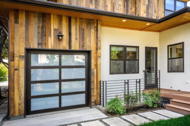 Modern residential front entry and garage facade featuring vertical reclaimed-looking timber cladding, a dark-framed sectional garage door with translucent panels, cream rendered walls, dark metal-framed windows, a timber soffit, covered porch, and horizontal metal railings. Concrete stepping slabs, planting, and lawn line the approach.