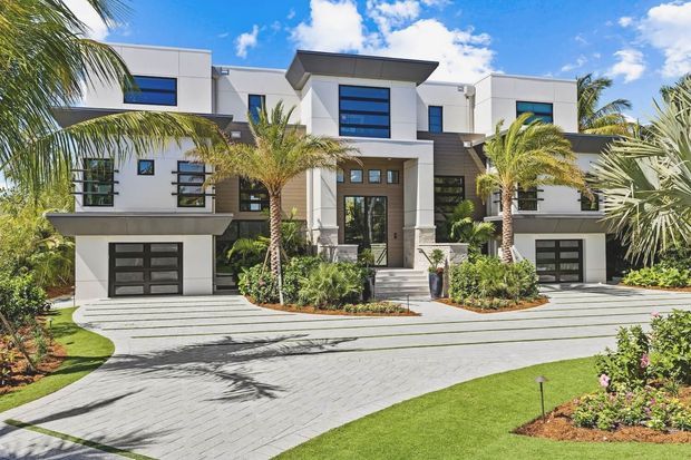 A large, symmetrical contemporary multi-storey residence viewed from the front, with crisp white rendered walls, dark horizontal cladding accents, flat projecting roof forms, extensive dark-framed glazing, and two integrated garages. A broad pale paved driveway leads to a formal central entry framed by columns and steps, with tropical palm landscaping and manicured lawns.