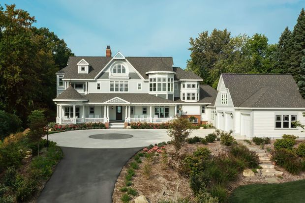 Exterior view of a large, traditional multi-gabled residence with white clapboard siding, dark shingle roofs, prominent dormers, ornate window groupings, covered verandas, and an attached garage. The home is surrounded by mature trees, landscaped planting, a curved paved driveway, and garden paths.
